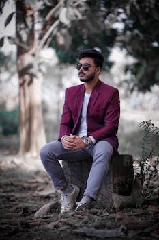 A fashionable man in sunglasses and a maroon suit sits outdoors on a tree trunk, exuding elegance.