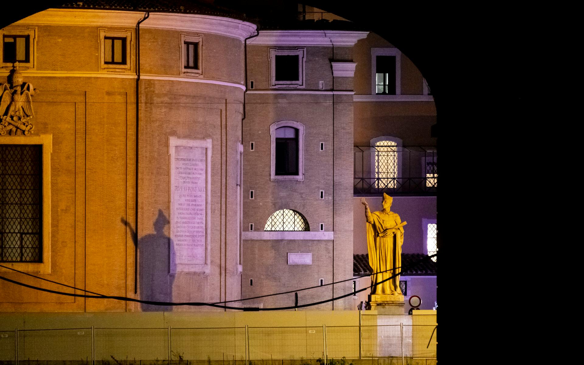 Illuminated statue casting a shadow against a building facade in Vatican City at night.