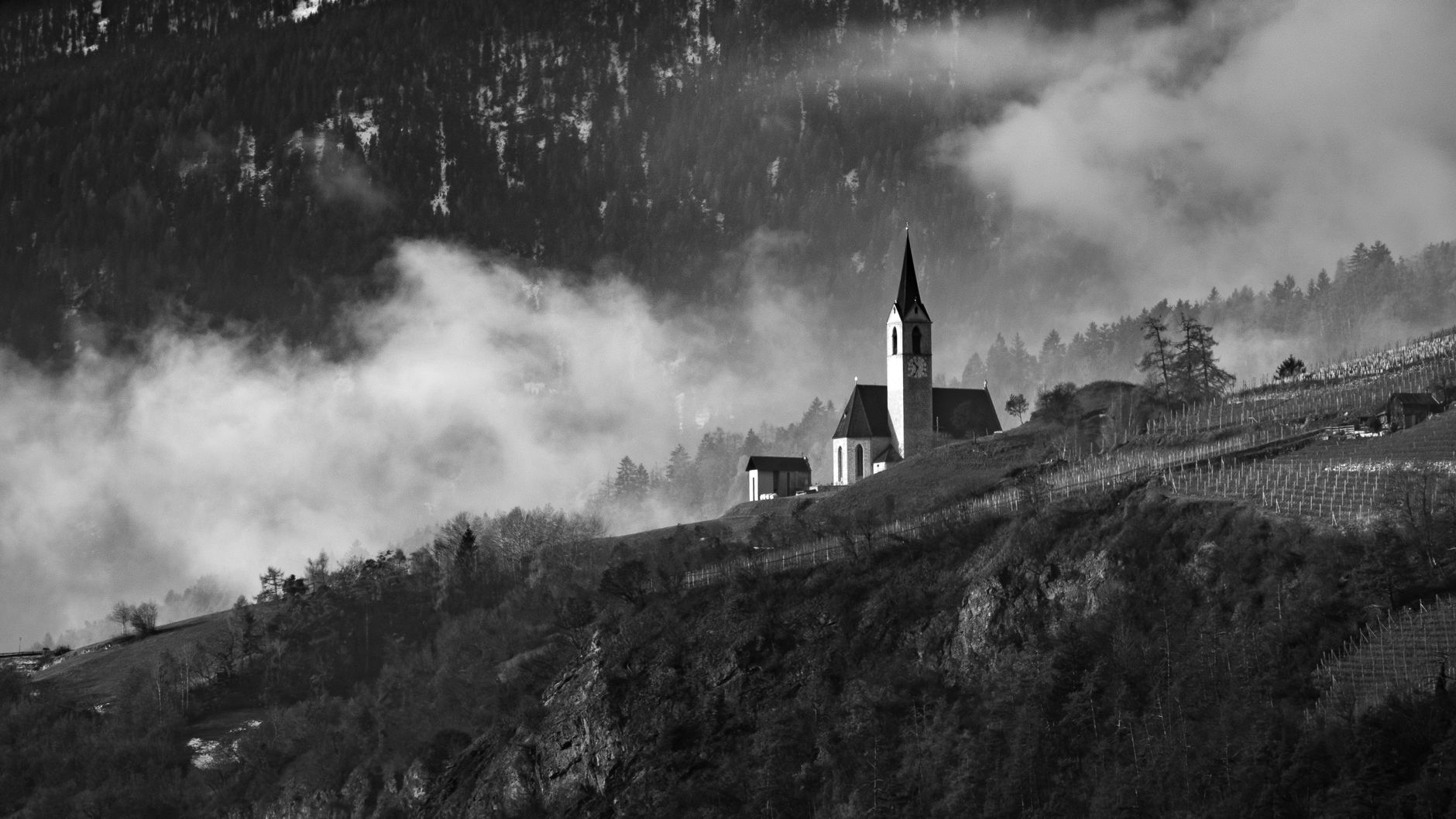 Black and white photo of a church on a misty hillside with mountains in the background.