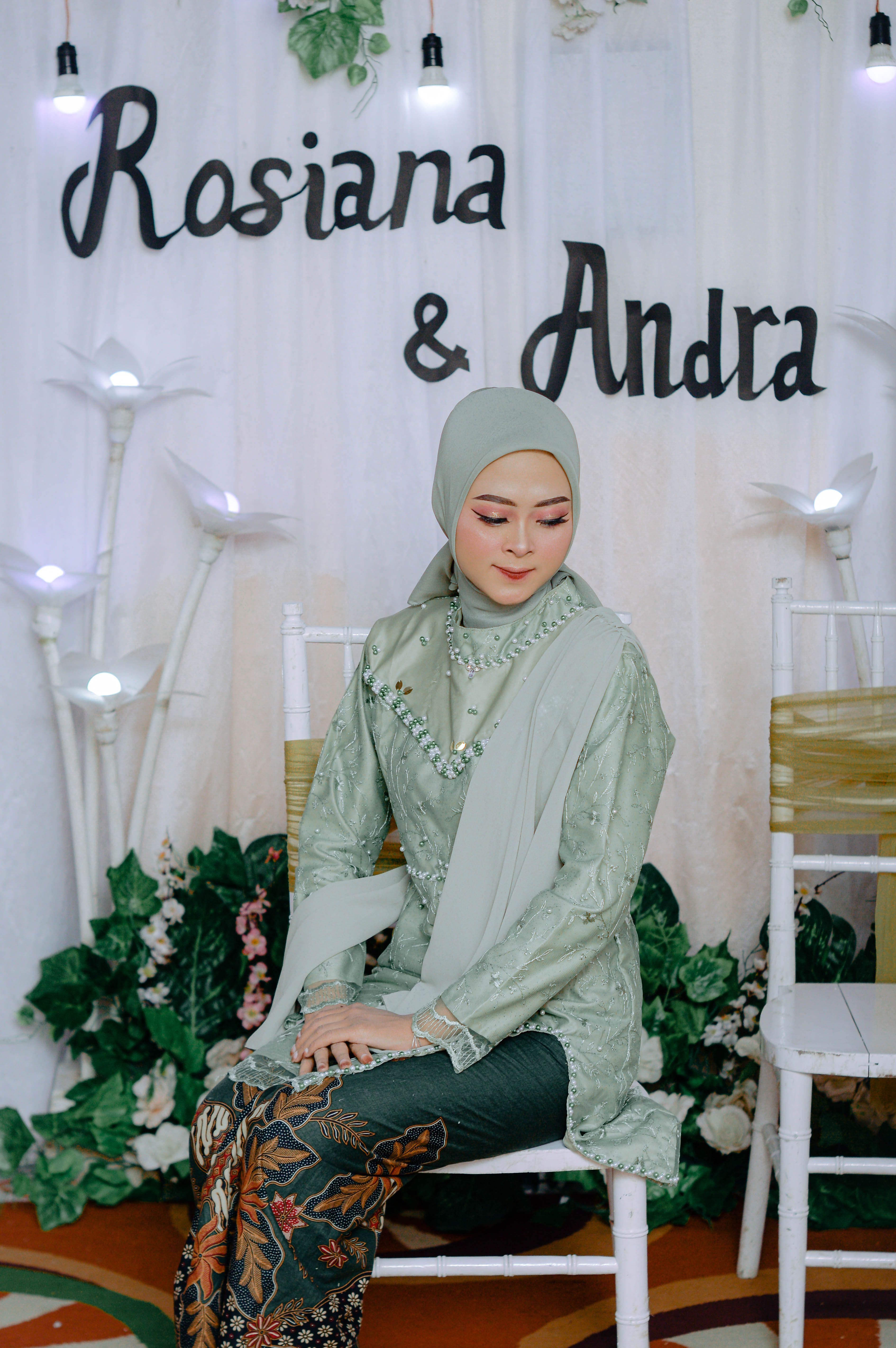 A woman wearing a hijab and traditional clothing sitting elegantly at a decorated event backdrop.
