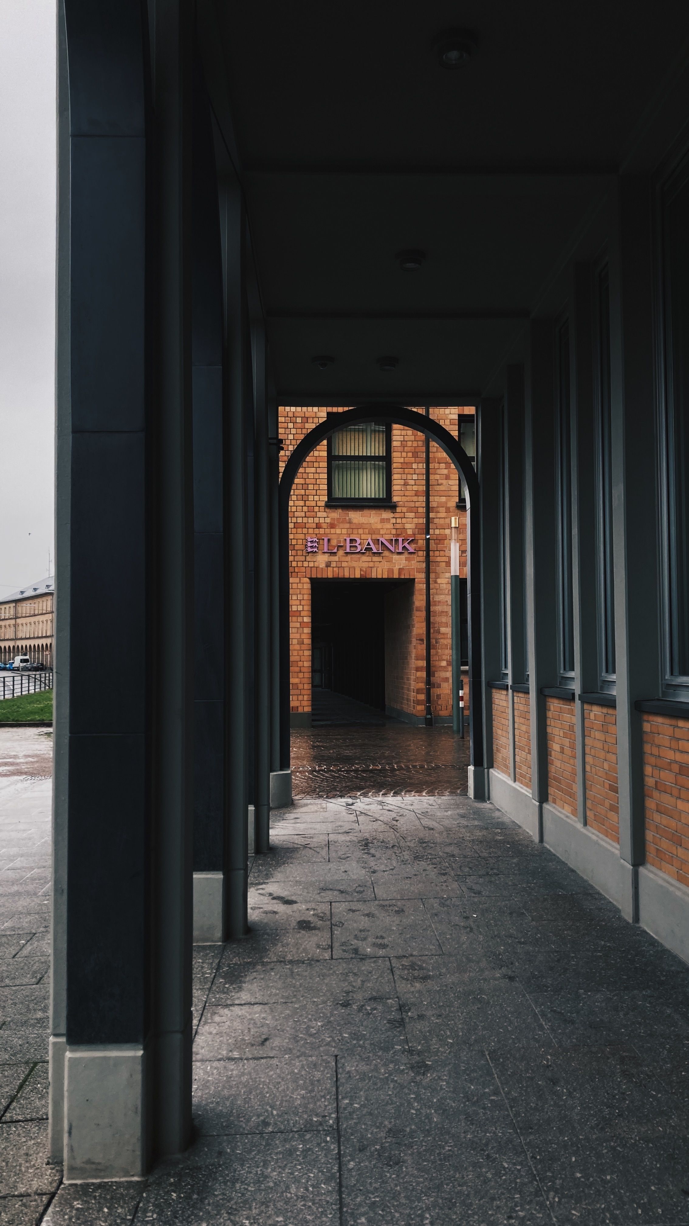 View through a modern colonnade to a brick bank entrance, creating a moody urban atmosphere.