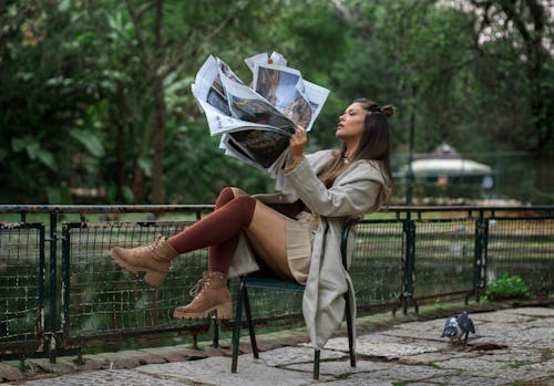 Young Woman Sitting on a Chair by the Pond and Holding a Newspaper 