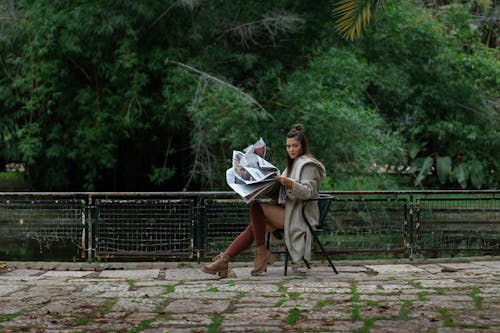 Woman Sitting on a Chair in a Park and Holding a Newspaper 