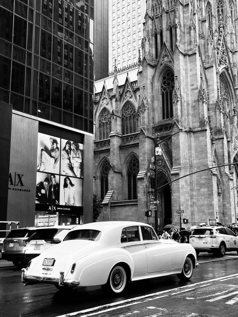 Black and white photo capturing a vintage car beside St. Patrick's Cathedral in bustling New York City.