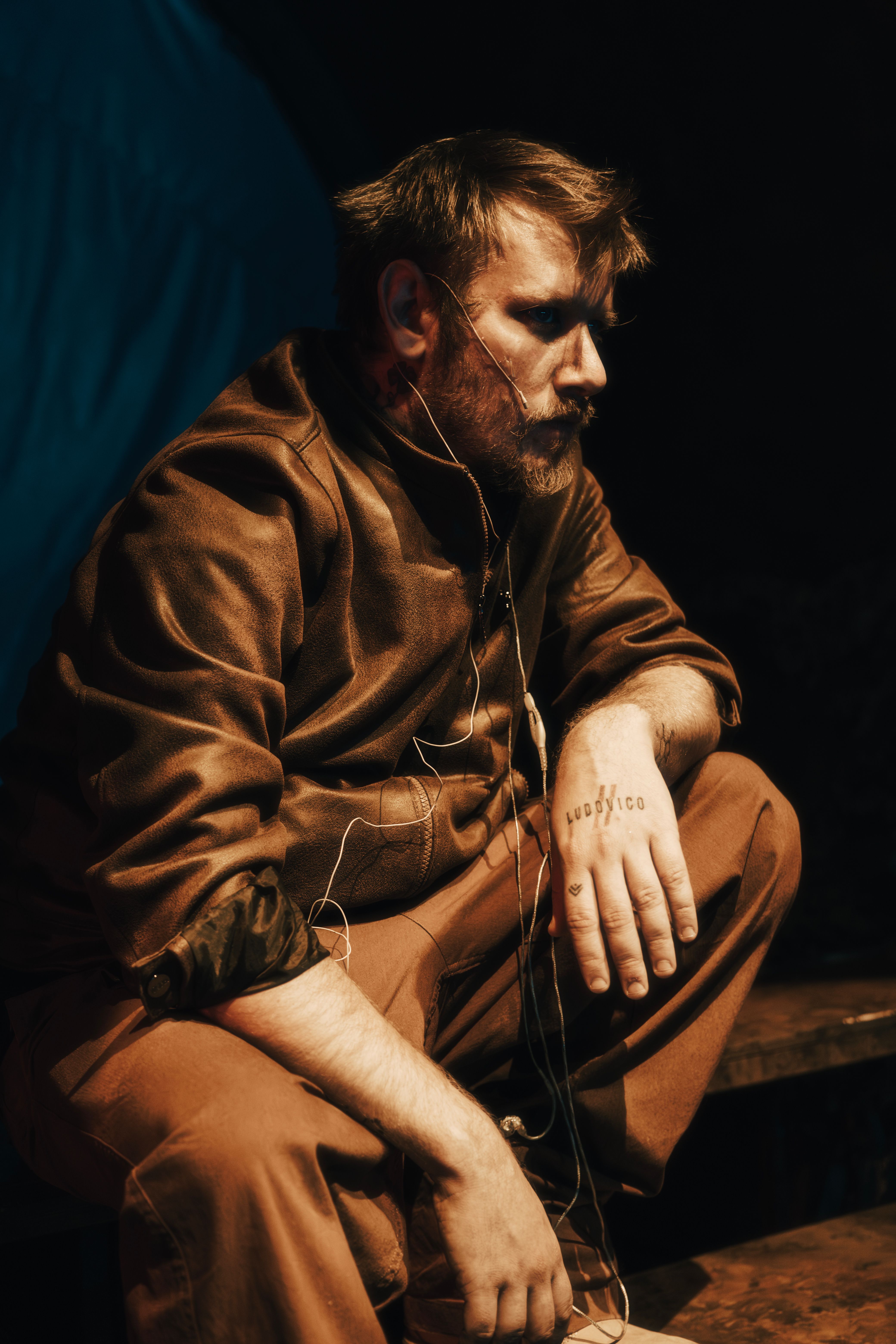 Man thoughtfully sitting indoors with a tattoo on his hand. Warm, moody lighting.