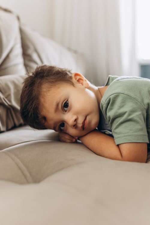 Free A young boy in a green shirt leans on a sofa, appearing thoughtful in an indoor setting. Stock Photo