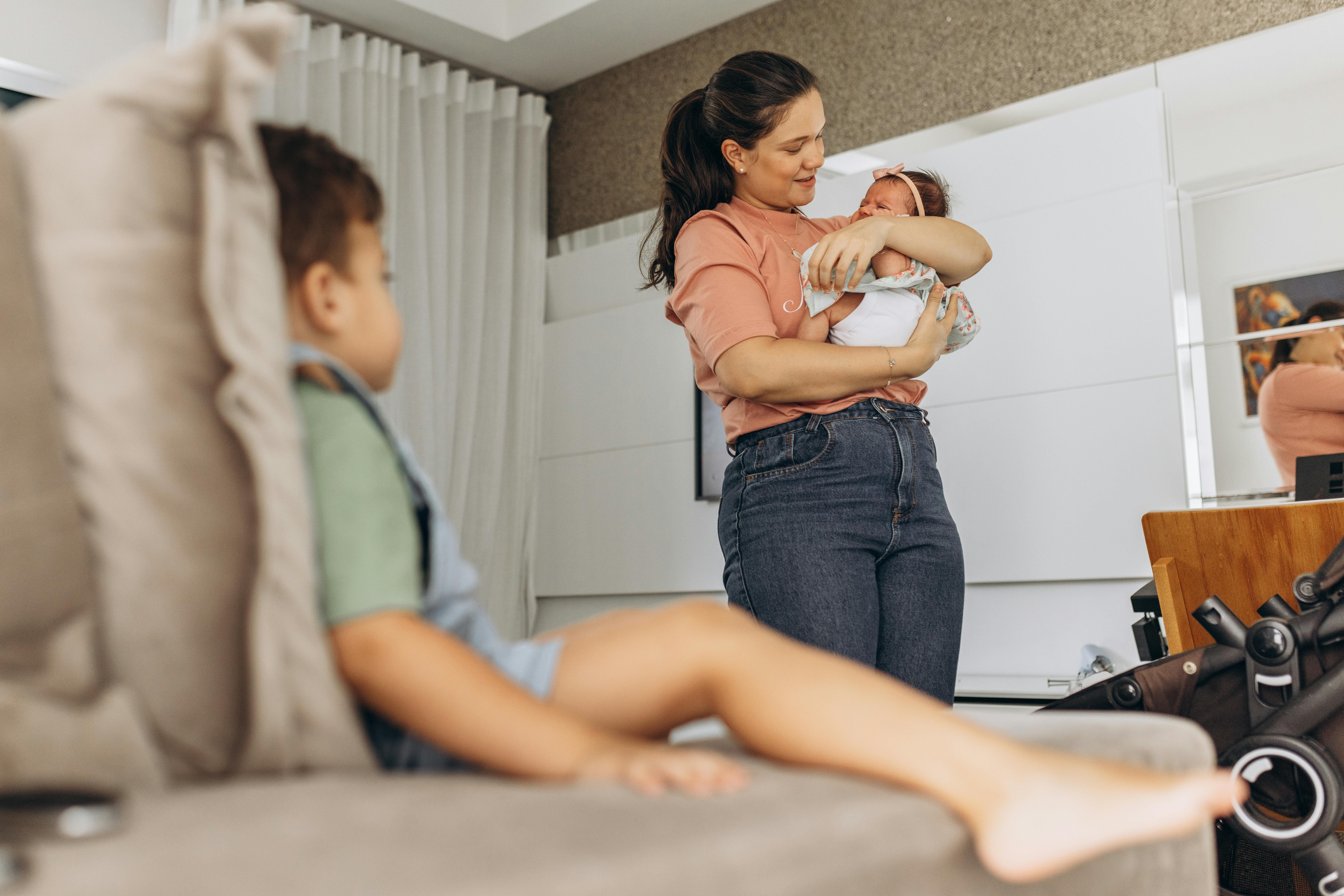 A mother lovingly holds her newborn while a toddler sits on the couch in a cozy living room setting.