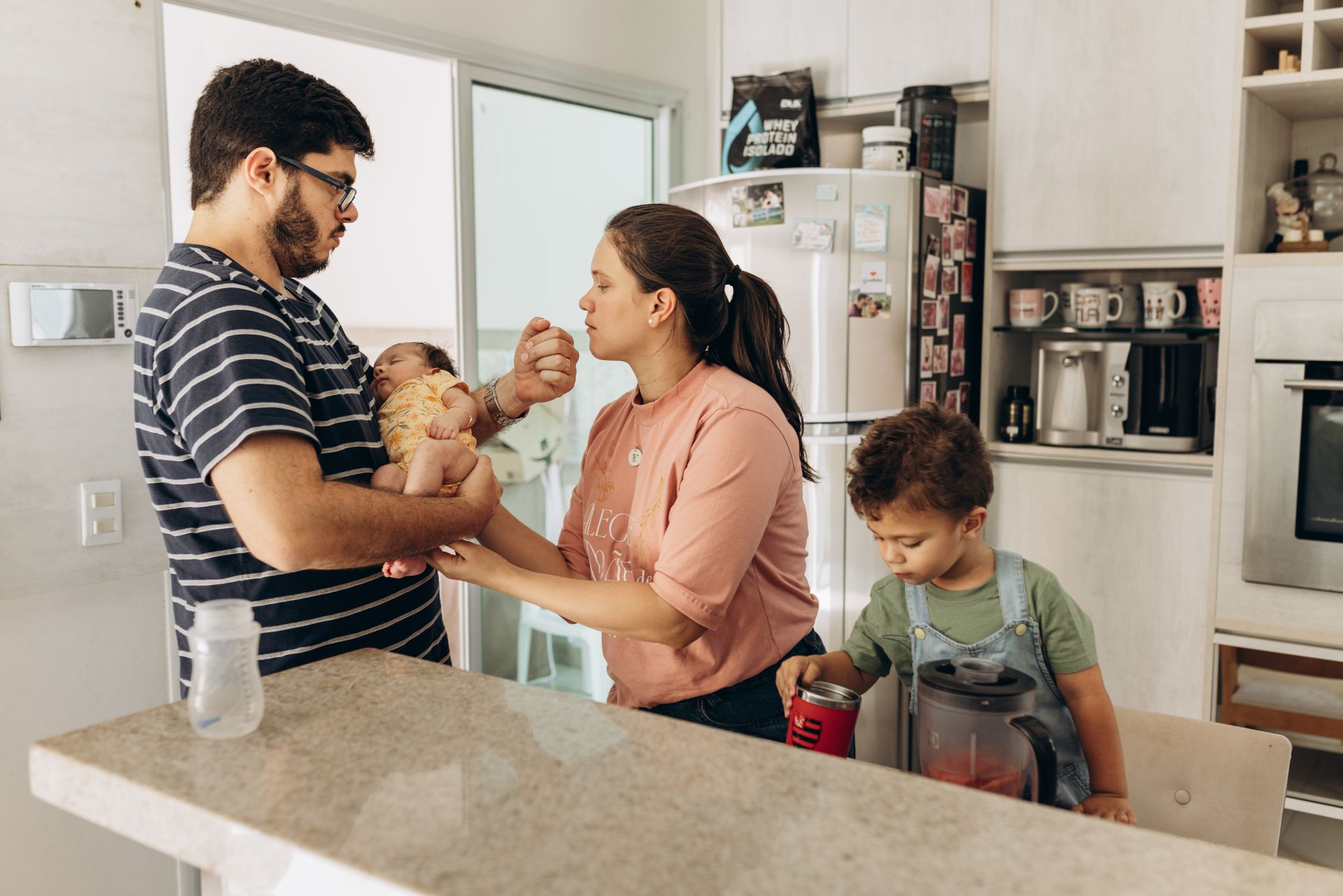 A family in a kitchen, with parents holding a newborn and a young child at the counter.