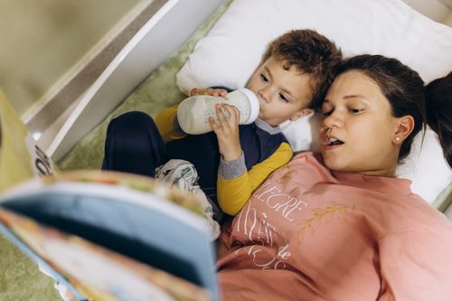 Free Mother and child lying in bed, bonding over a bedtime story and bottle feeding. Stock Photo