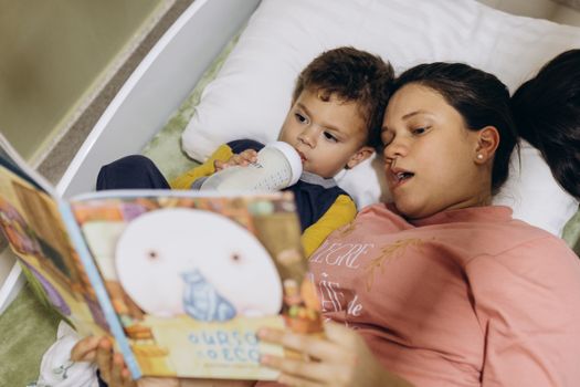 A mother reads a bedtime story to her child while he drinks milk, creating a cozy and bonding moment.