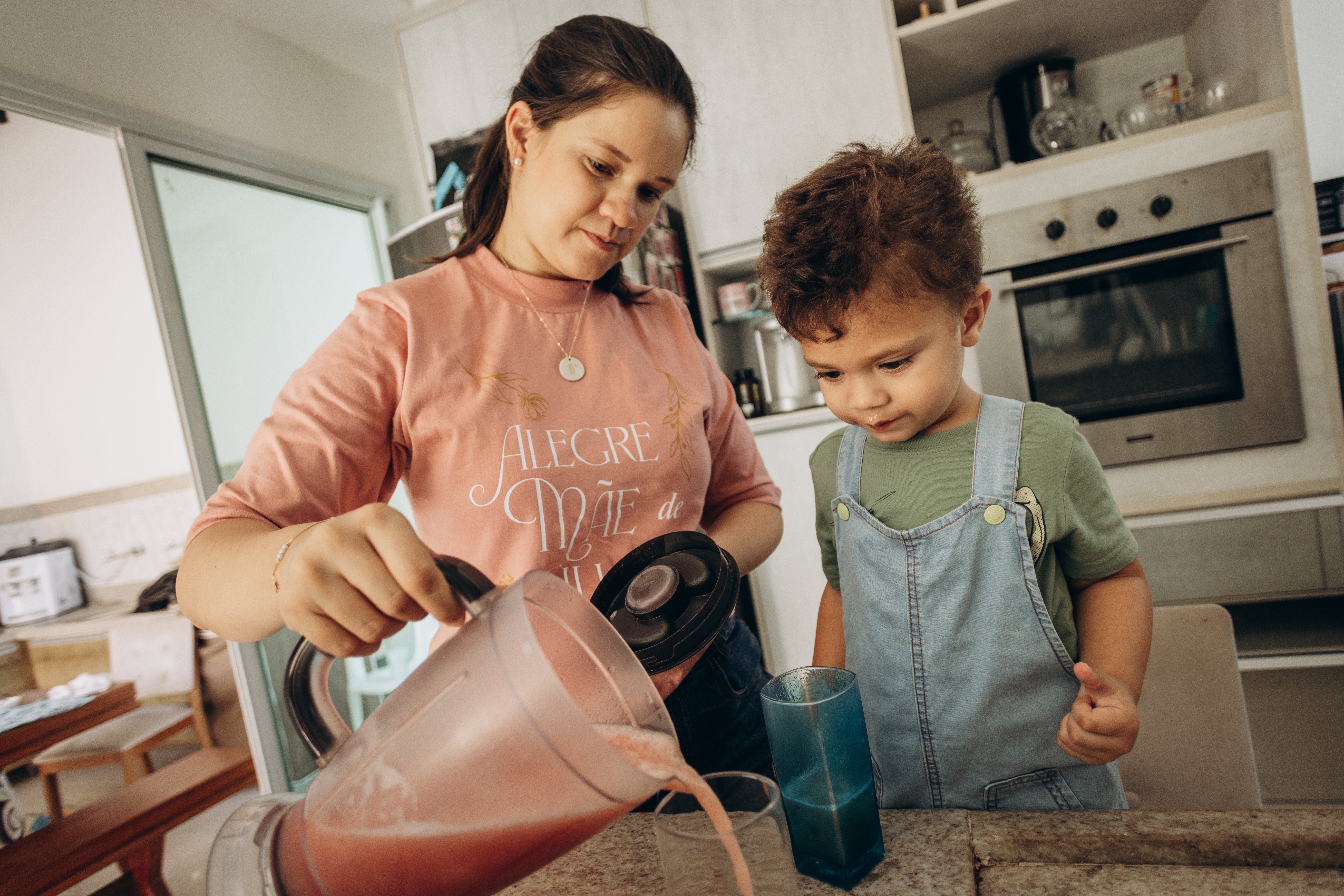 A mother pours a smoothie for her son in a home kitchen, showcasing family bonding.
