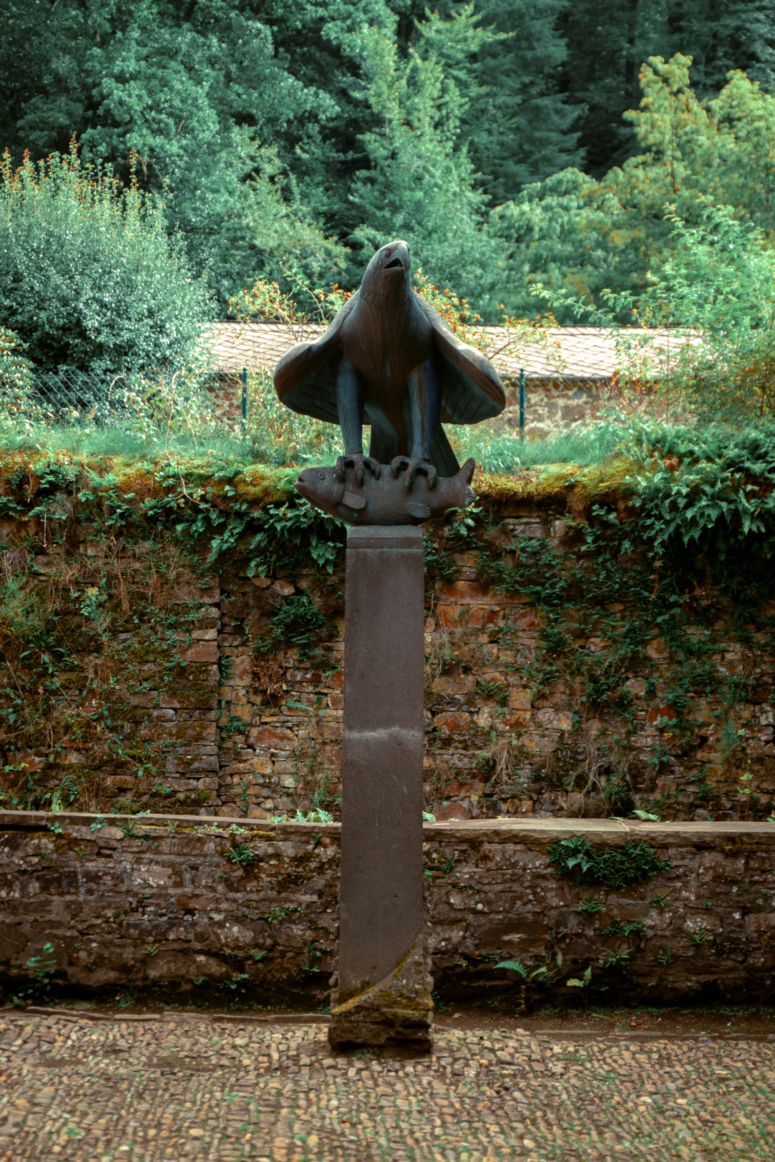 An artistic eagle sculpture on a pedestal in a lush garden setting.