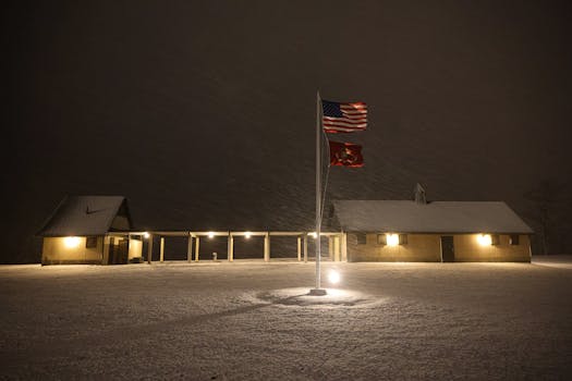 Warm lights glow in snowy storm around houses and flagpole at night.