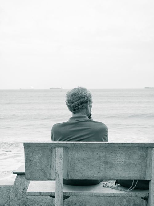 Free A man sits alone on a bench facing the calm sea, lost in thought. Stock Photo