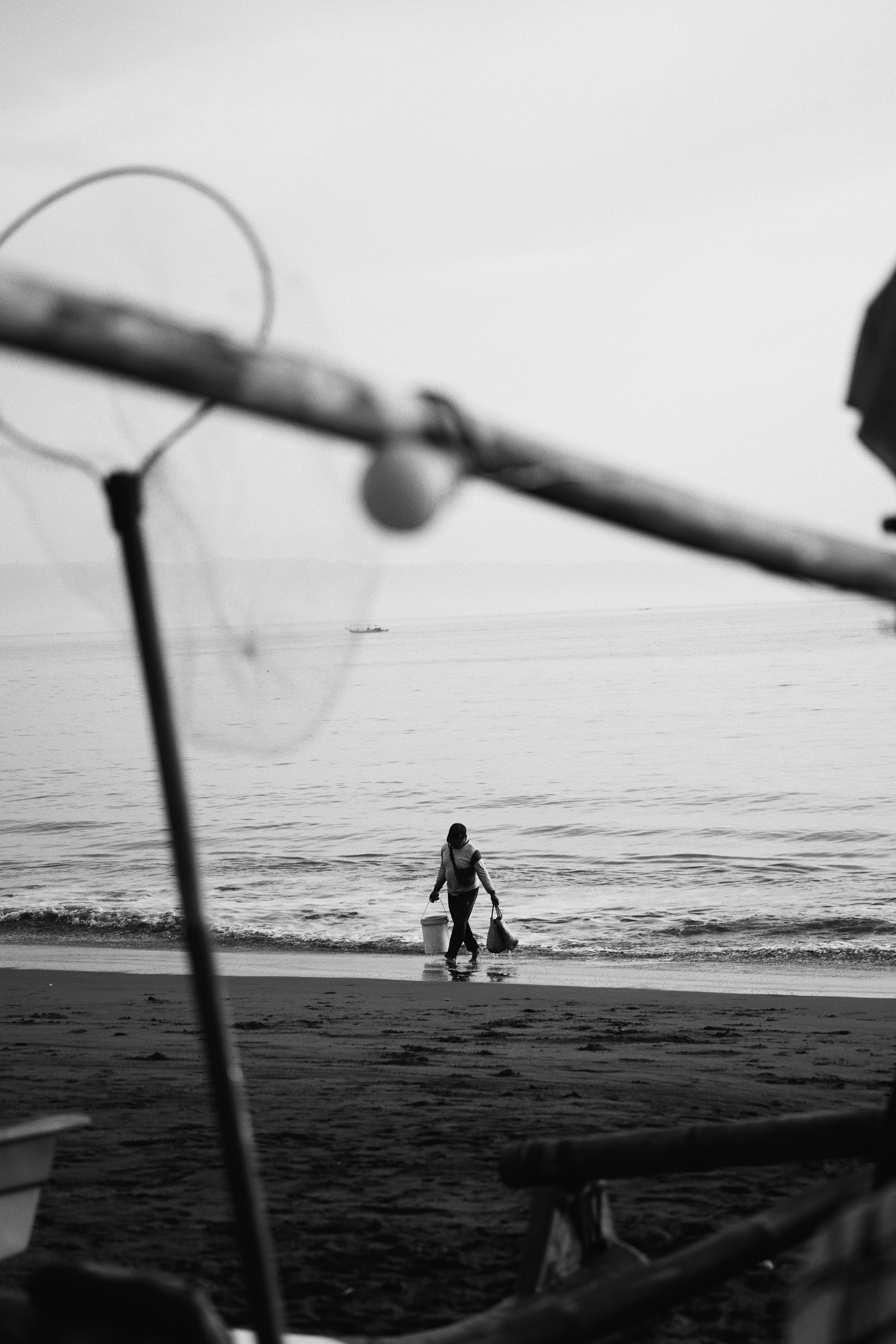 A fisherman carrying buckets along a quiet beach, captured in black and white.