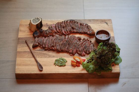 Sliced grilled flank steak on a wooden board with a roasted marrow bone, sauces and lettuce