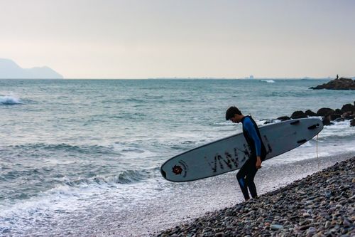Free A lone surfer carrying a surfboard walks along a rocky beach in Lima, Peru, at sunset. Stock Photo
