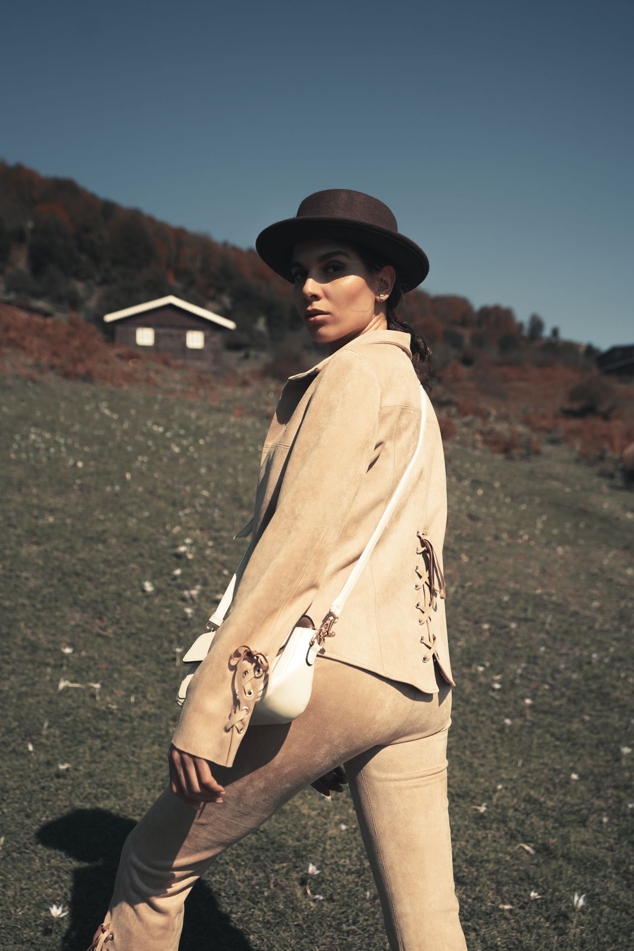 Woman in a matching beige suede jacket and pants set with a brown hat and white crossbody bag in a sunny meadow