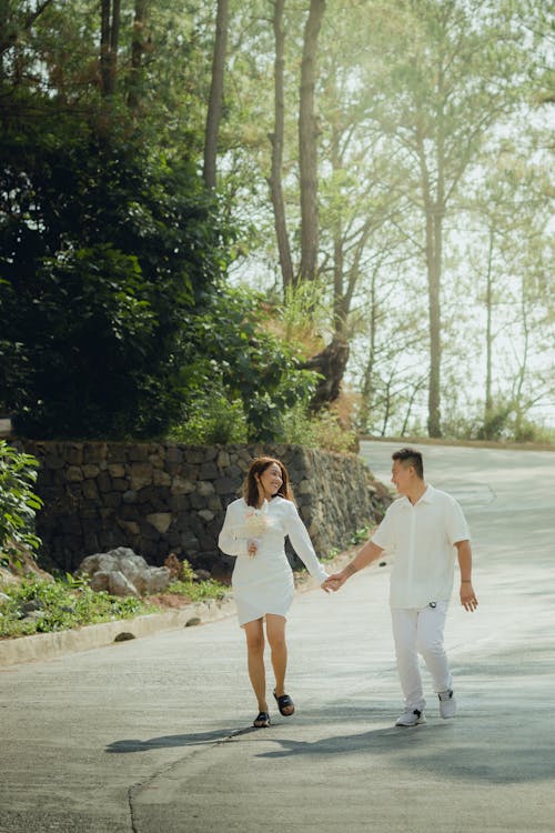 Woman in White Dress and Man in White Shirt Holding Hands and Walking on Road