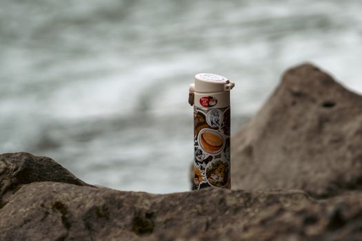 A vacuum flask decorated with stickers rests on rocks by a seashore in Russia.