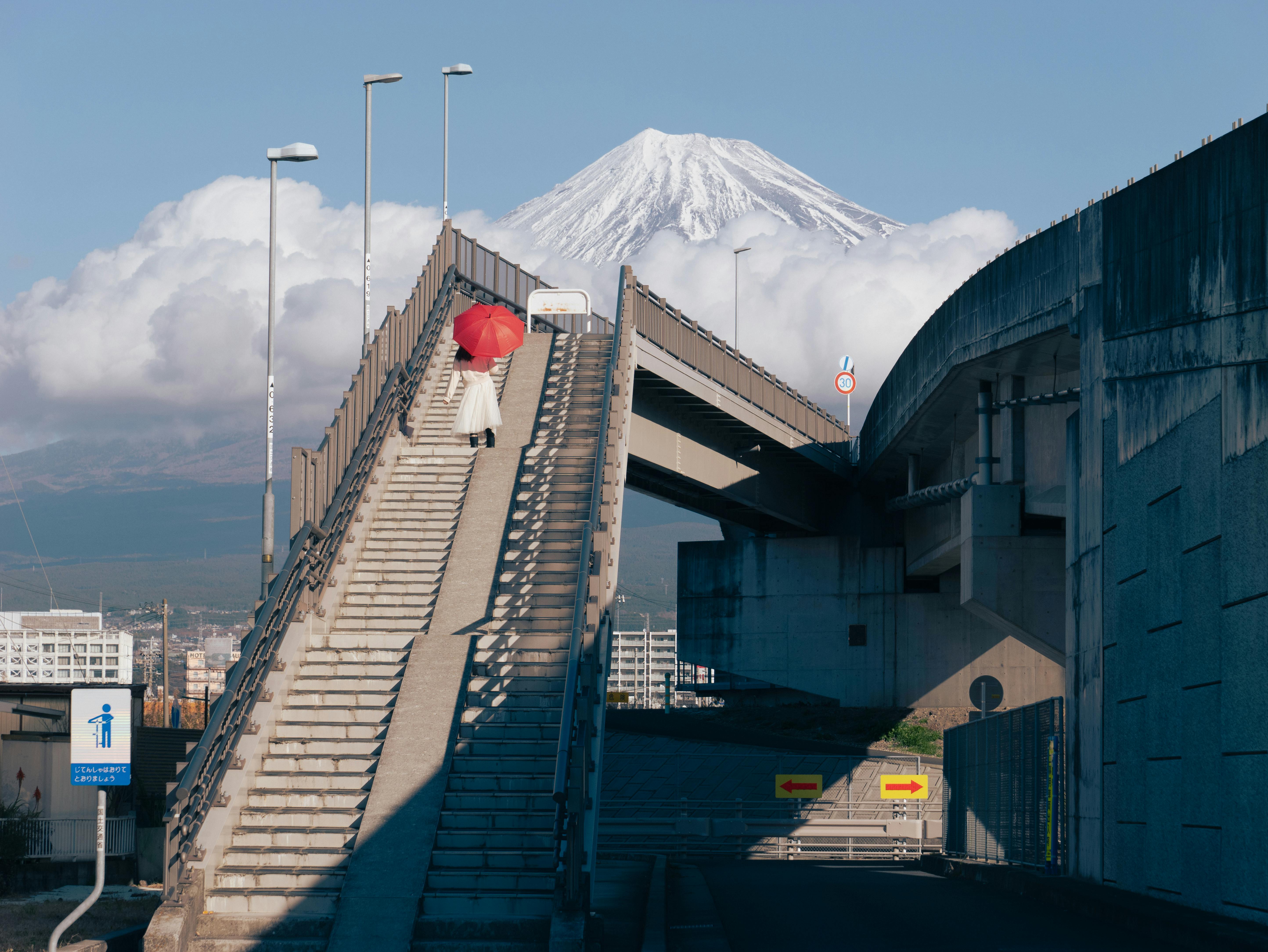A woman with a red umbrella walks up steps with Mount Fuji in the background, captured in Japan.