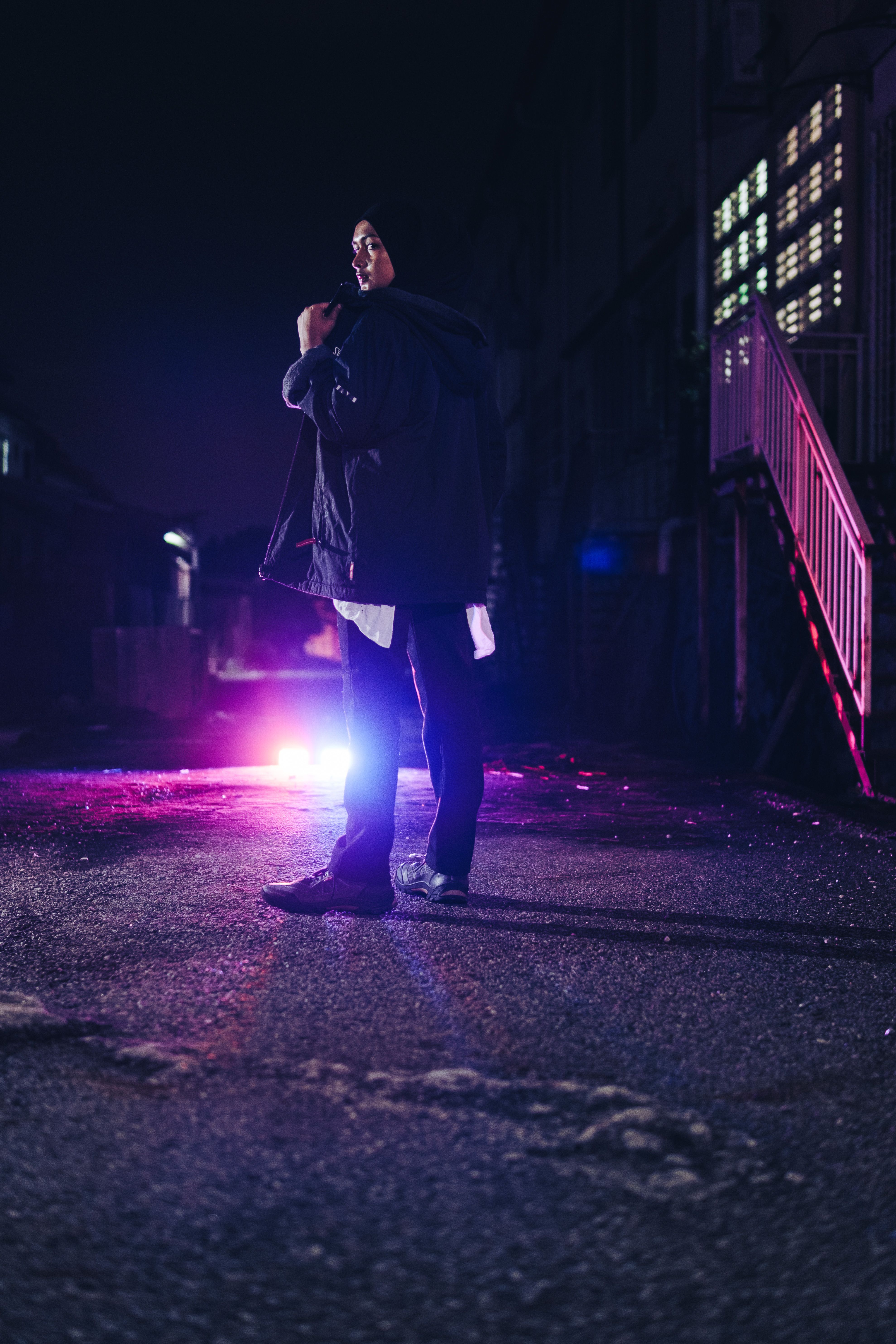 Portrait of a young woman in hijab with jacket, standing on city street at night with purple lights.