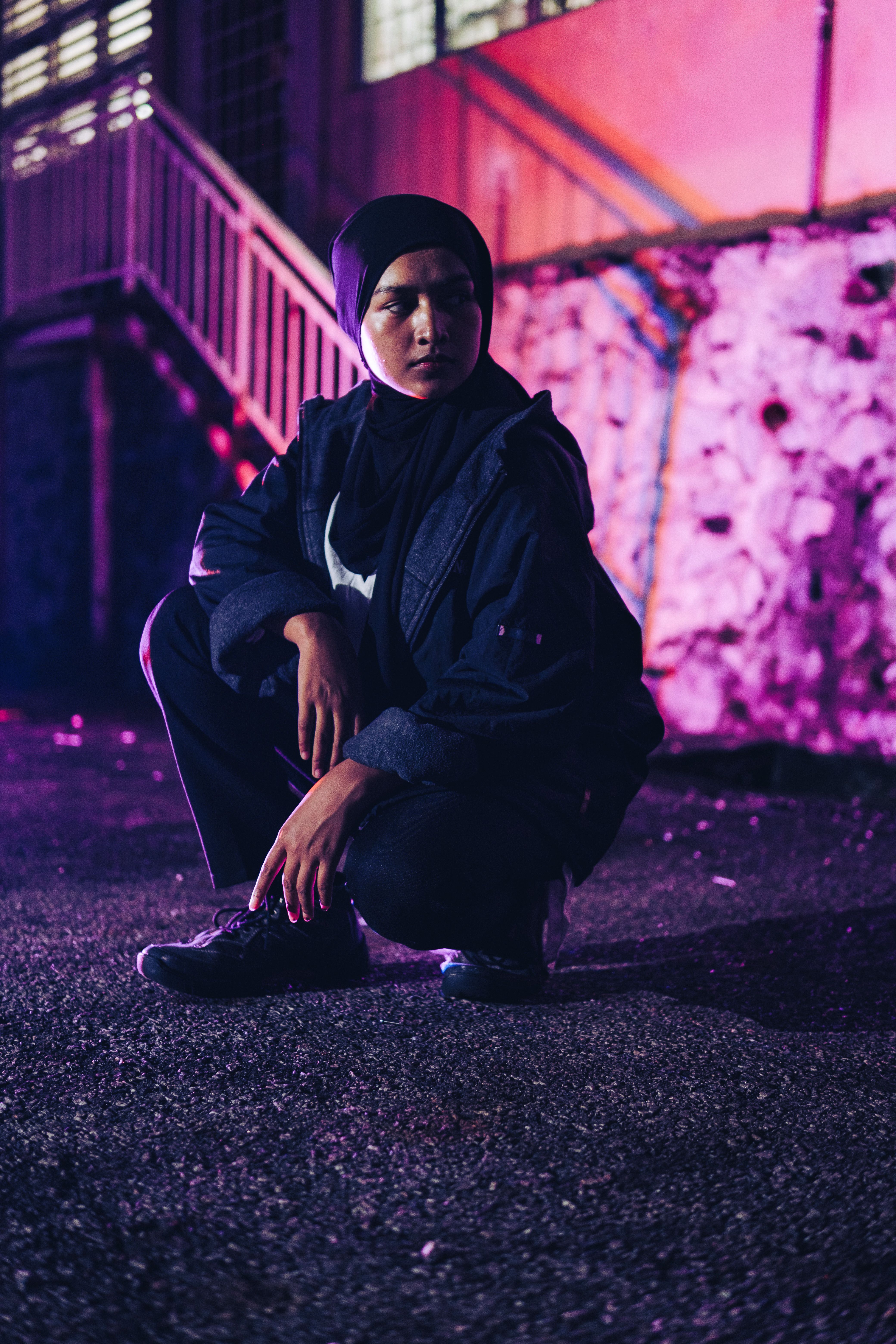 Stylish portrait of a young woman wearing a hijab, crouching on city streets under vibrant purple lights at night.