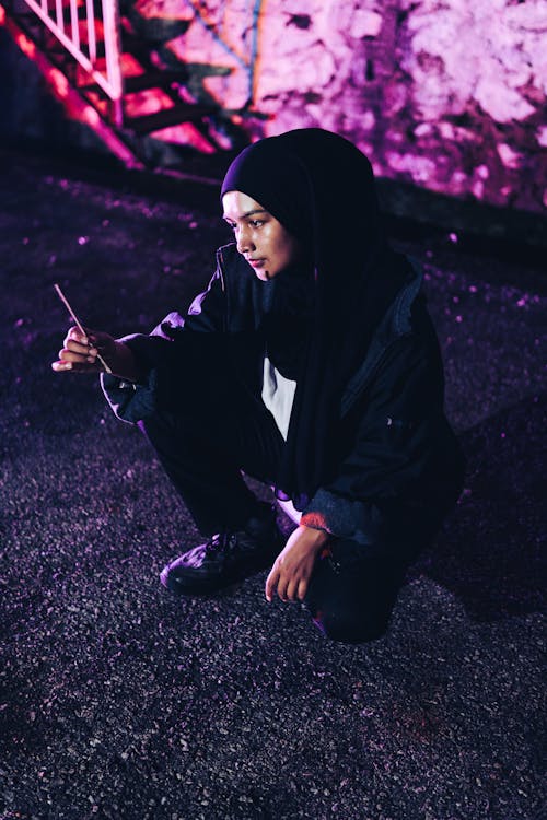 Portrait of a Female Model Wearing a Hijab Crouching Outdoors with a Smartphone in Hand