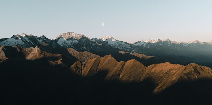 A breathtaking aerial view of a snowcapped mountain range with clear skies and a crescent moon.