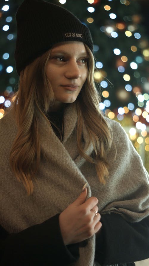 Young Woman in a Hat and Scarf Standing Outside on the Background of Lights 