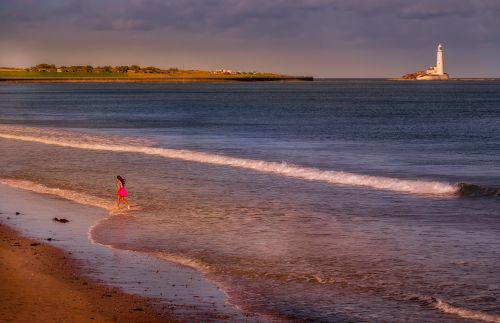 Free A young girl runs along the shore near a lighthouse during sunset, with waves gently crashing. Stock Photo