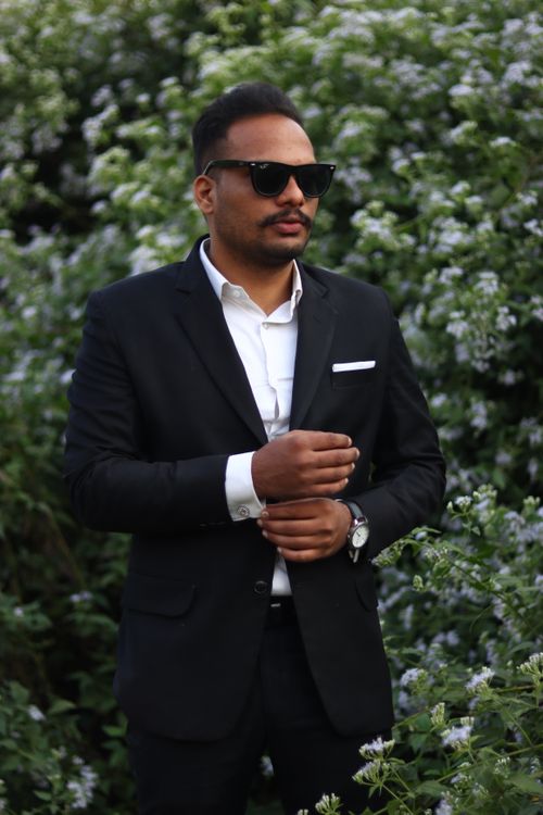 Free A fashionable man stands confidently in a black suit surrounded by lush greenery and white flowers. Stock Photo