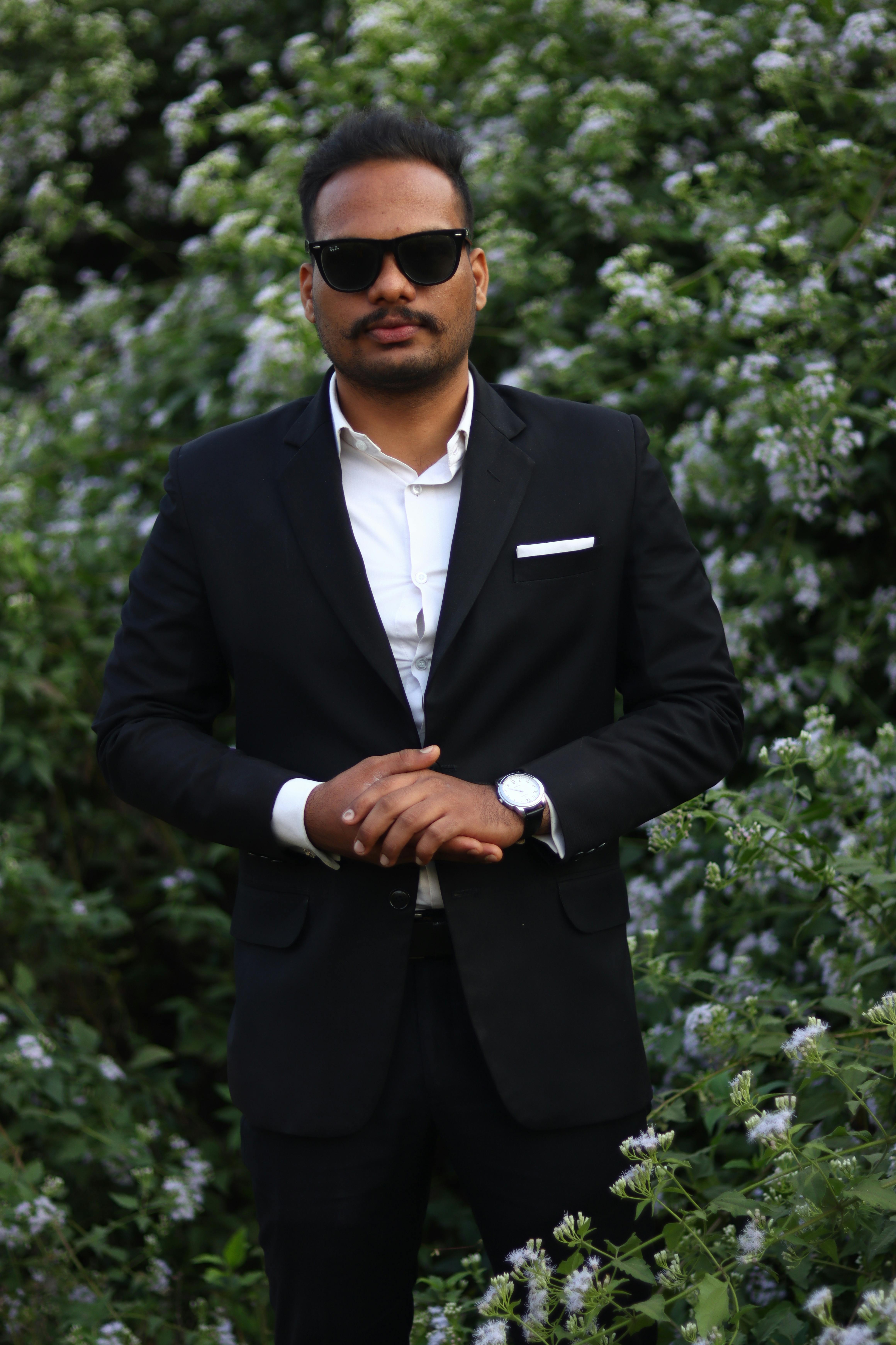Man in black suit posing confidently amidst lush flower garden.