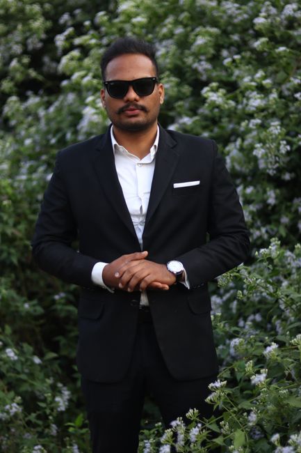Man in black suit posing confidently amidst lush flower garden.