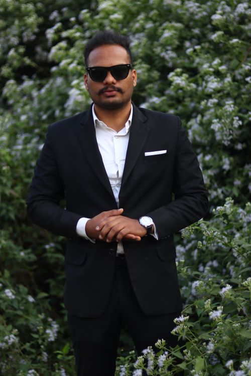 Free Man in black suit posing confidently amidst lush flower garden. Stock Photo