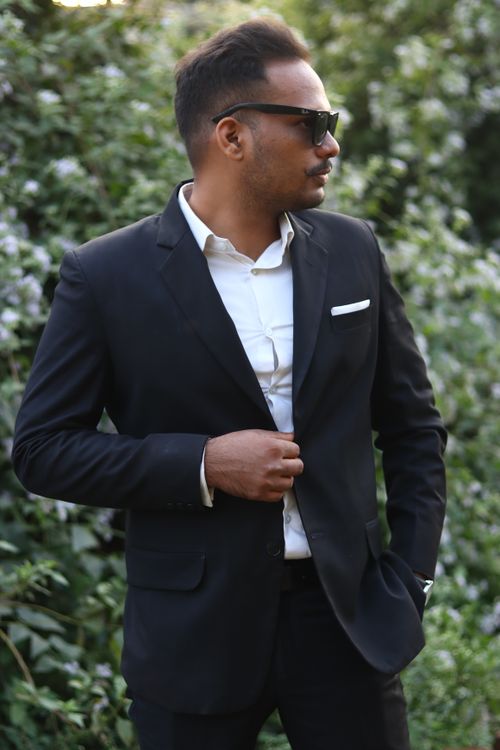 Free Elegant man in a black suit and sunglasses posing in front of a flower bush outdoors. Stock Photo