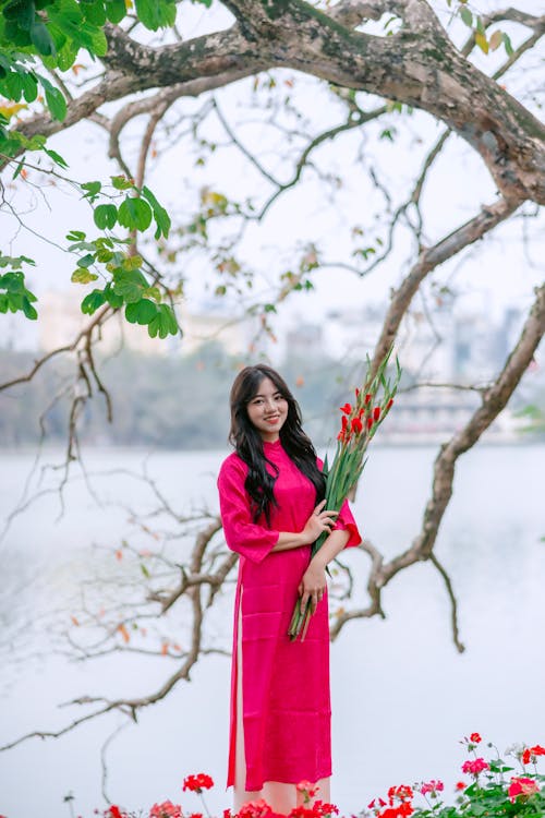 Young Model in Pink Ao Dai Dress by the River Holding Flowers