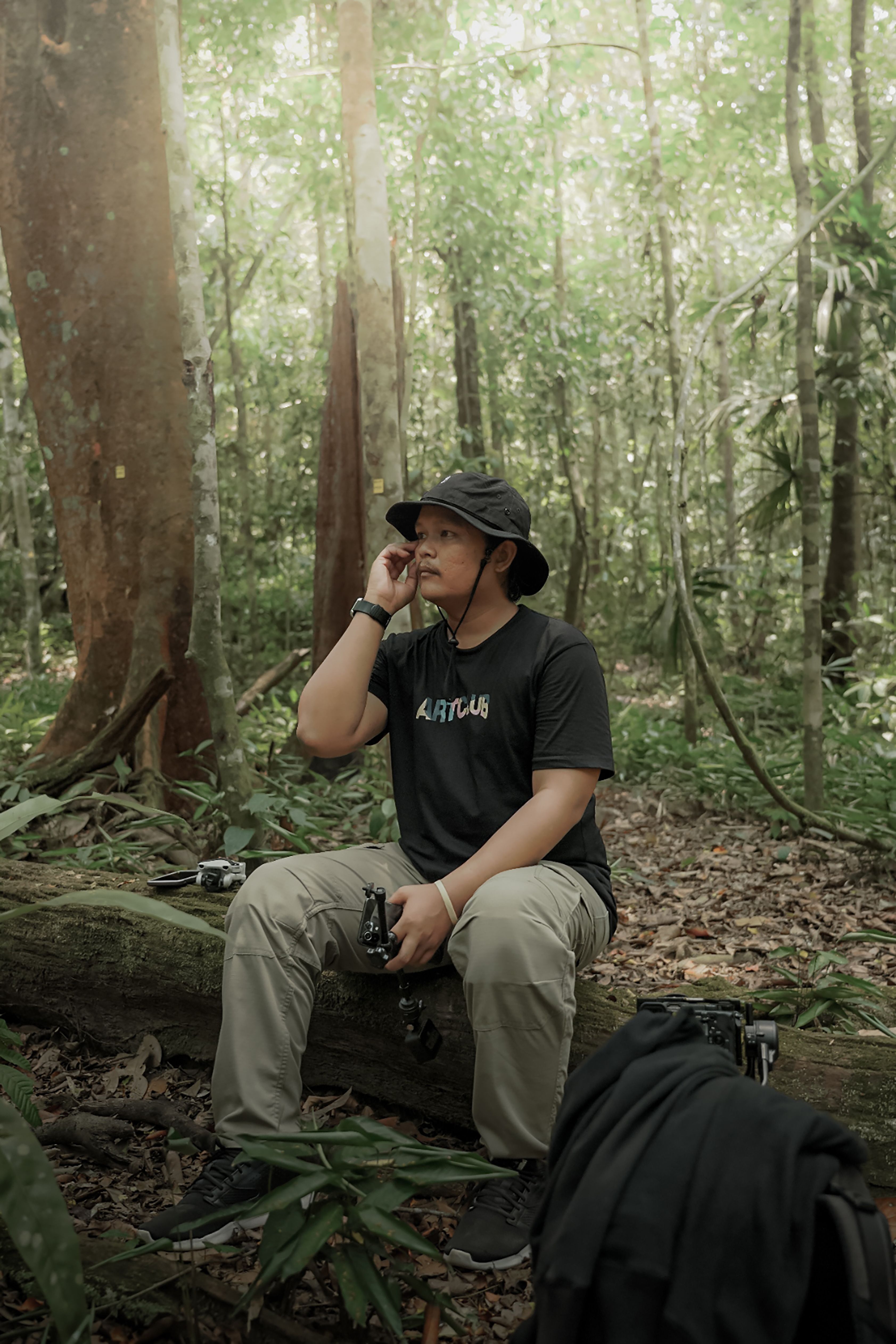 Man in a forest, wearing a hat, sitting on a log with a camera, enjoying nature.