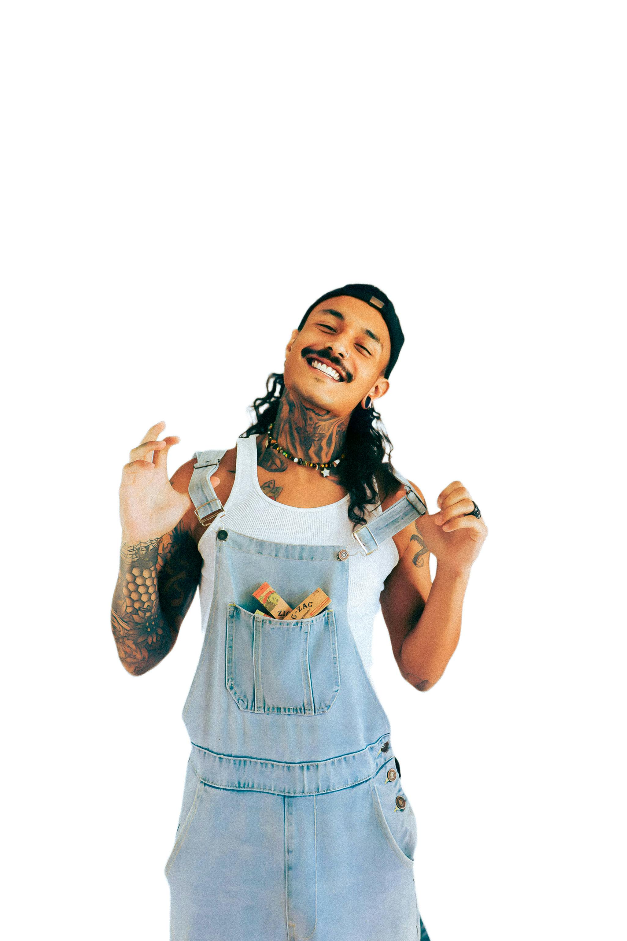 Smiling tattooed man in denim overalls with casual pose in studio setting.