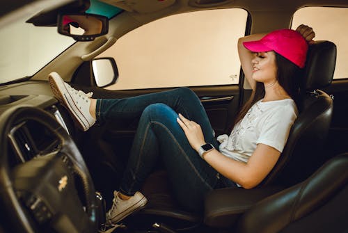 Woman in a car wearing a pink cap and jeans, smiling and relaxed.