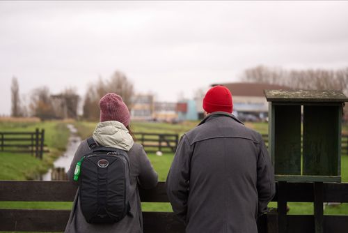 Free A couple dressed warmly observes a scenic farm landscape from behind a fence, suggesting leisure and travel. Stock Photo