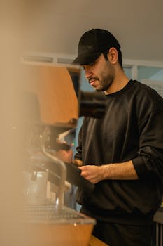 A barista wearing a cap operates a coffee machine in a busy Ankara café.