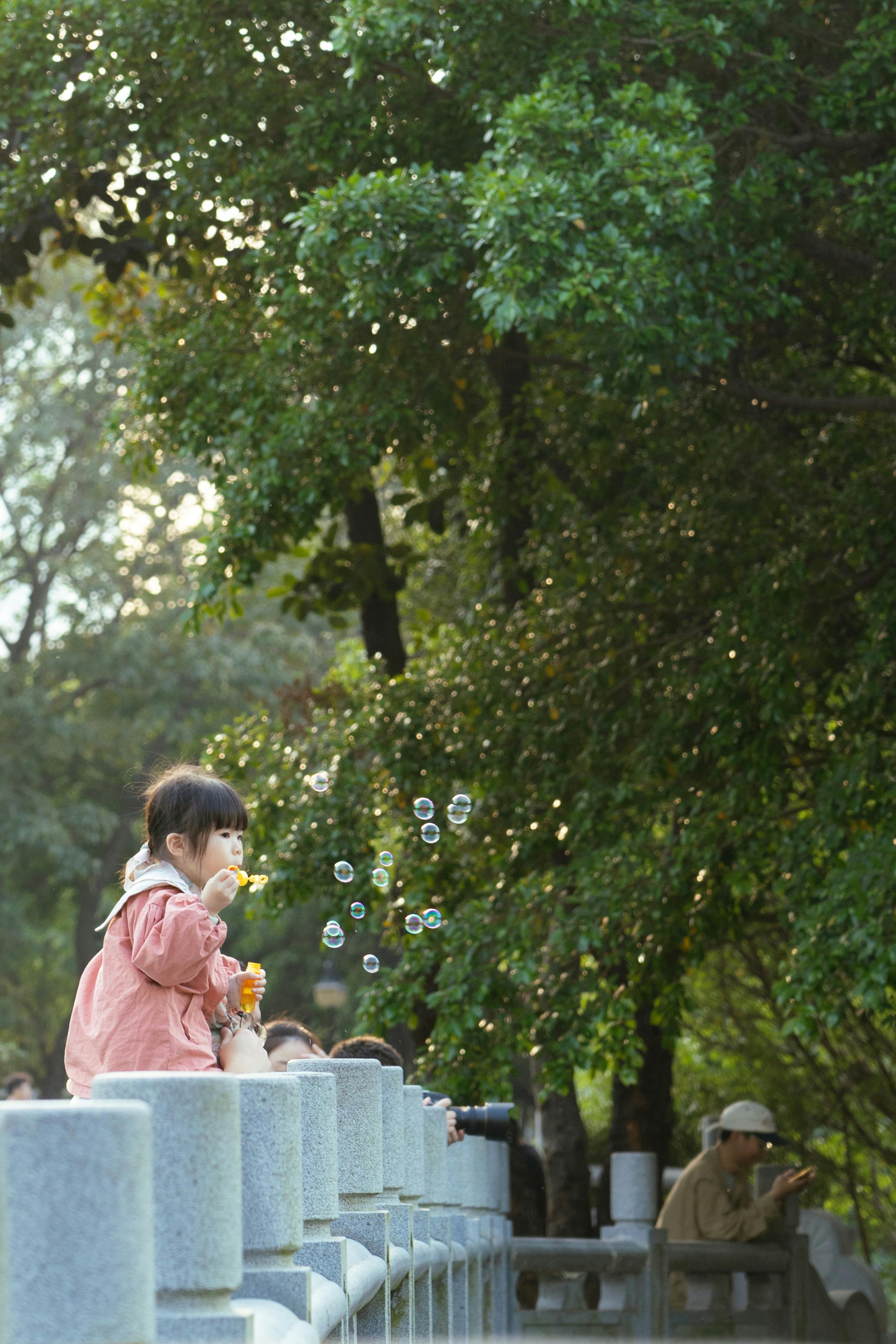 A young girl enjoys blowing bubbles on a sunny day in a verdant park, capturing a moment of pure joy.