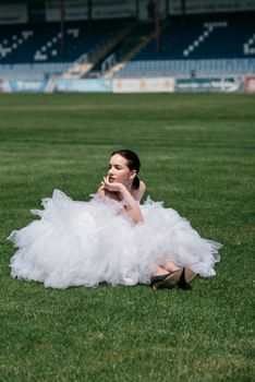 A woman in a white dress sits on a green sports field under the sun.