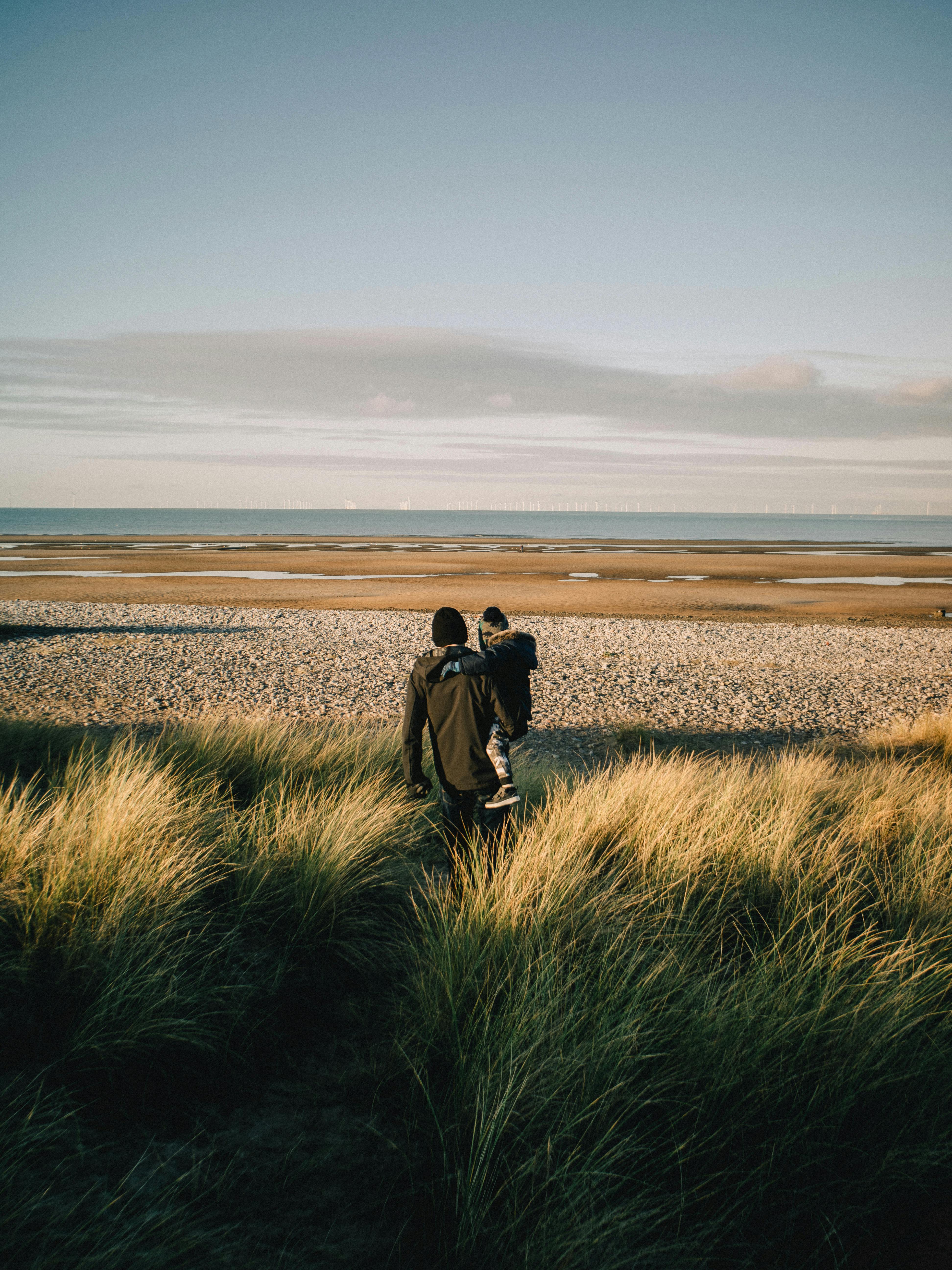 A parent and child walk along a sandy beach under a clear sky, dressed for cool weather.