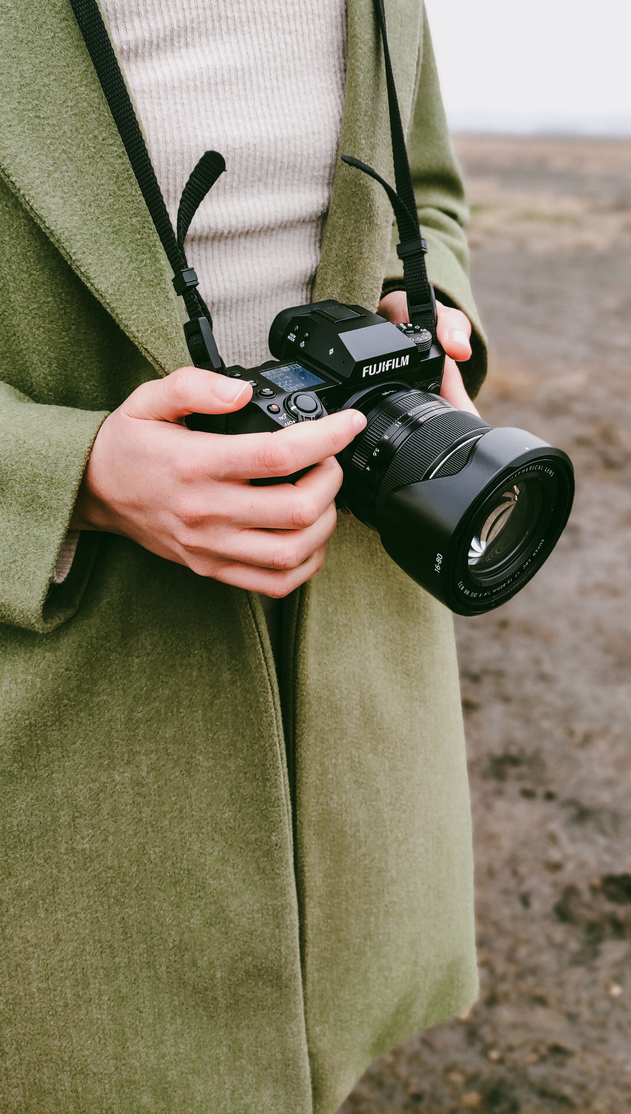 A person in a green coat holding a camera outdoors on a cloudy day.