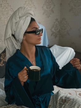 A woman in a bathrobe with a towel on her head enjoys coffee indoors, embracing a calming morning routine.