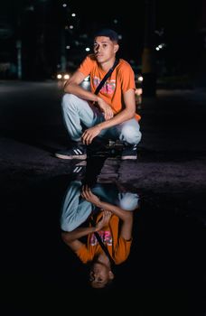 A teenager in street wear squats by a puddle at night, with a vivid reflection.