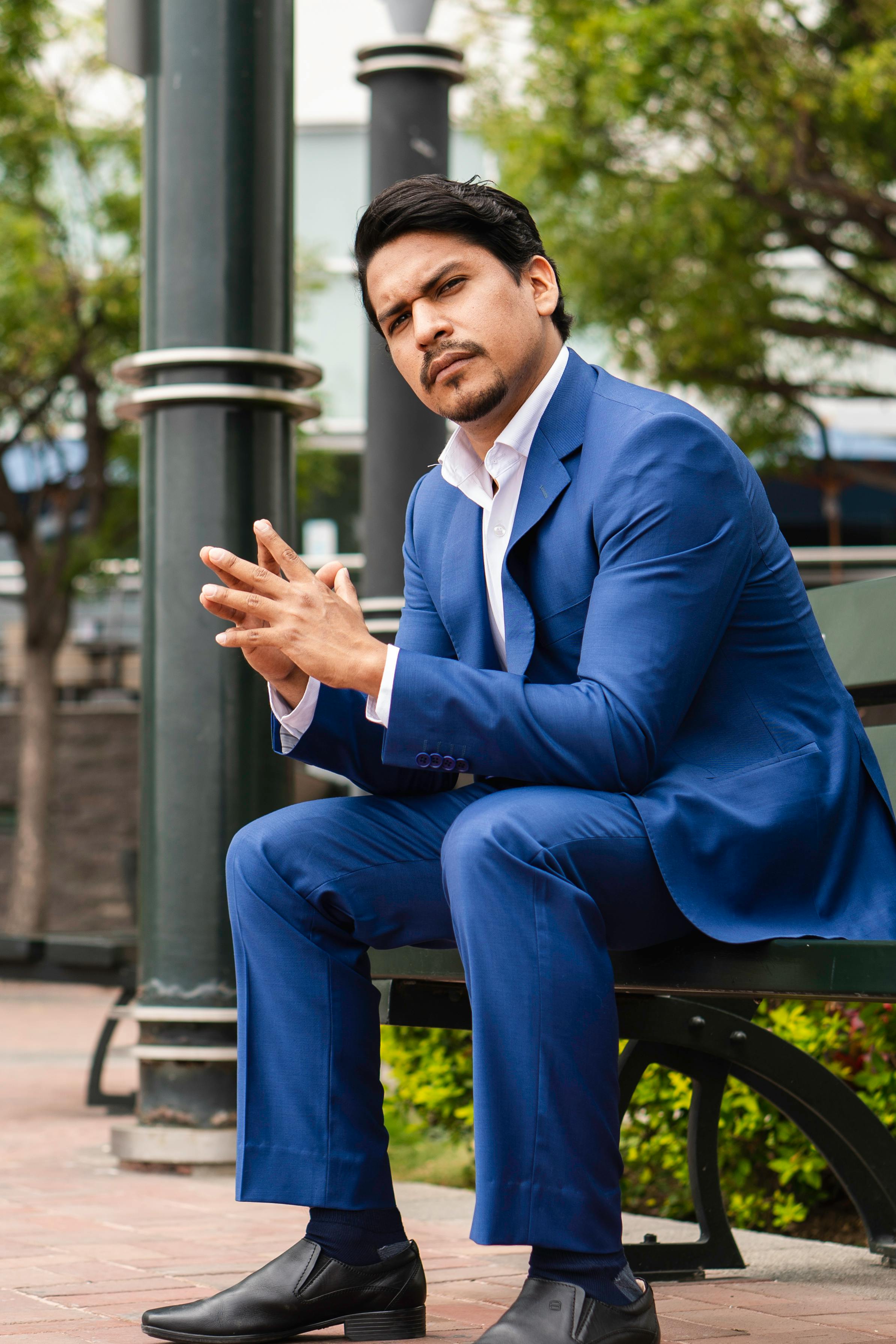 Businessman in a blue suit sitting on a bench in an urban park setting, showcasing elegance and confidence.