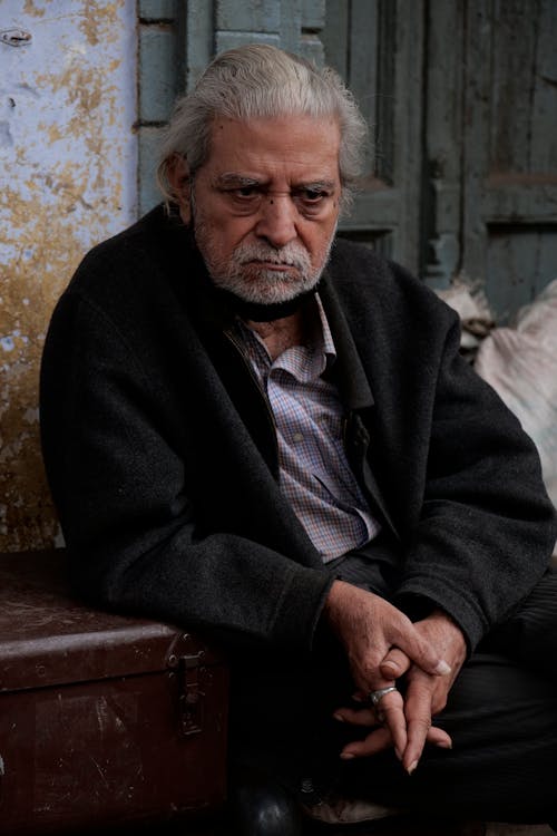 Photo of an Elderly Man with Gray Hair and Beard Sitting by the Wall 